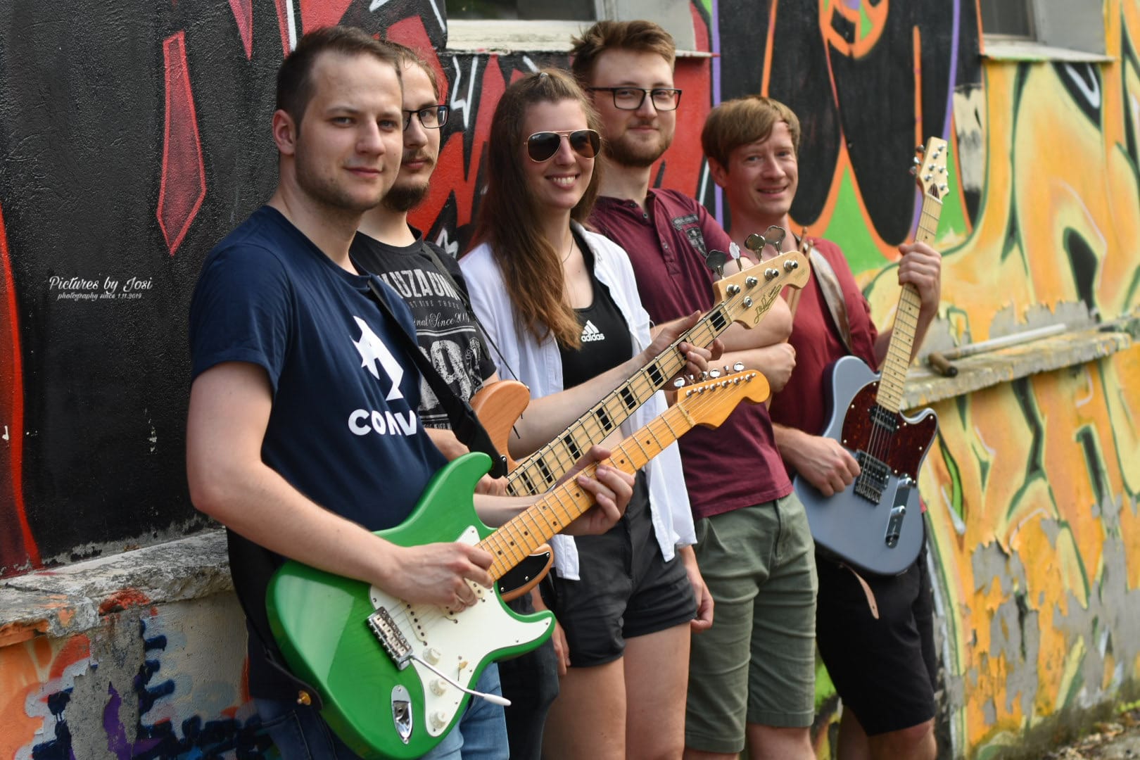 Zu sehen ist die Band Air Shot. Die fünf Bandmitglieder stehen vor einer mit Graffiti besprühten Wand. Drei der Personen halten E-Gitarren bzw. einen E-Bass in der Hand.